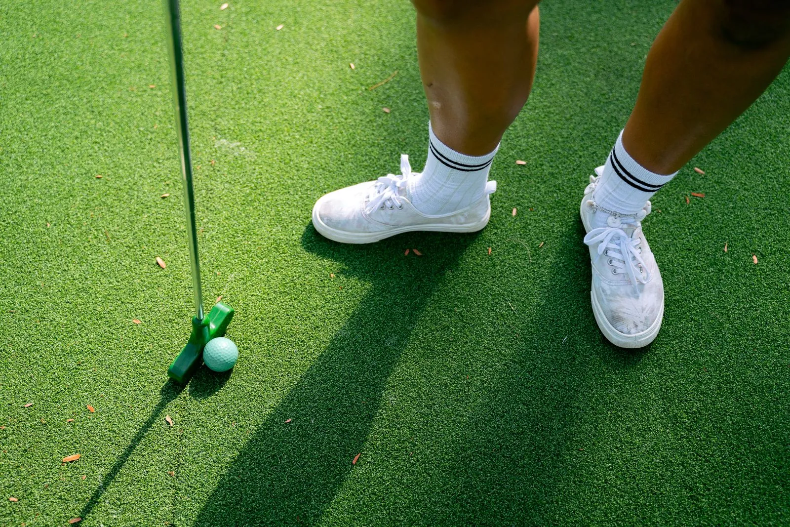 A person in white sneakers standing with a putter and golf ball on a green putting surface.